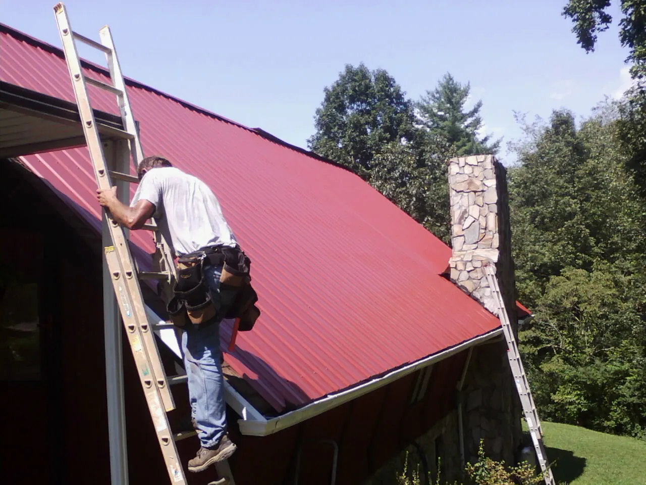 Roofer on a ladder beside a red metal roof and stone chimney — Soffit & Fascia Repair by Roofing Brothers in East Saint Louis